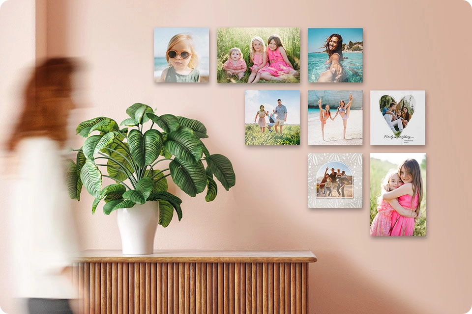 Woman walks by a set of eight Snapfish Photo Tiles featuring family photos hung on a wall above a table with a houseplant